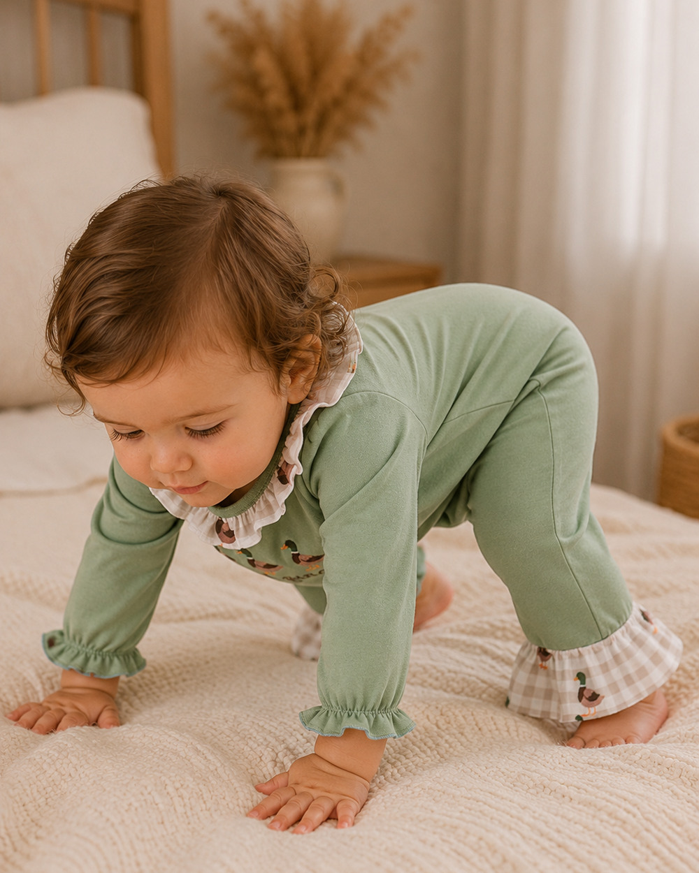 A baby crawling on a cream-colored textured bed, wearing a sage green long-sleeve jumpsuit with ruffled trim, gingham cuffs, and small embroidered details.