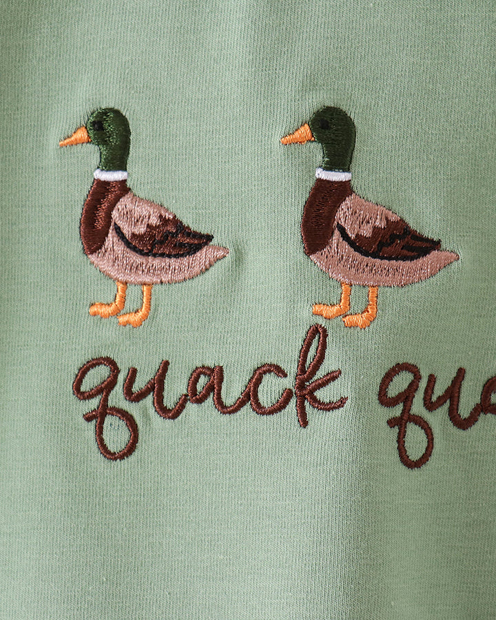 Close-up of a sage green fabric item featuring two embroidered mallards with orange beaks and feet, above the cursive text &