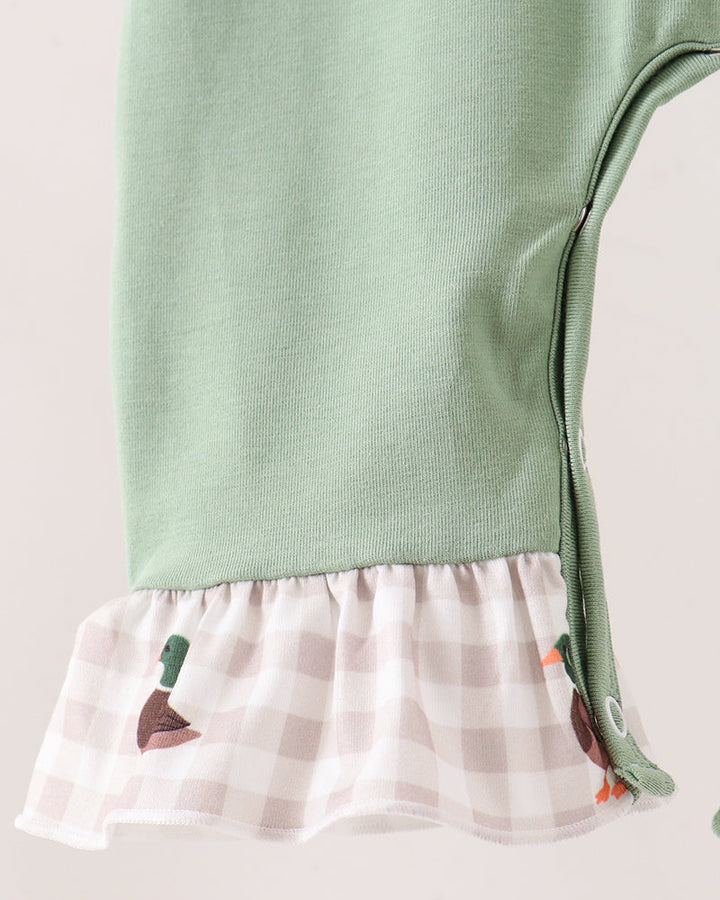 Close-up of a baby garment with a sage green upper section and a ruffled hem made of white and pale pink gingham fabric with small duck and carrot motifs.