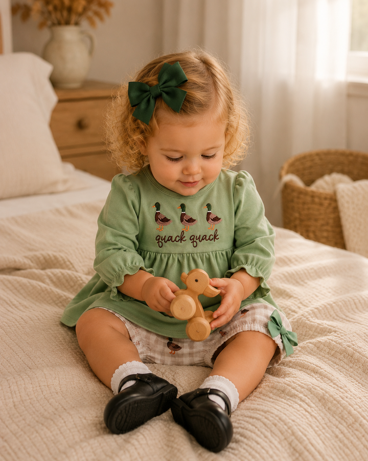 A toddler girl sits on a cream-colored bed, wearing a sage green long-sleeve dress with embroidered ducks and the text &