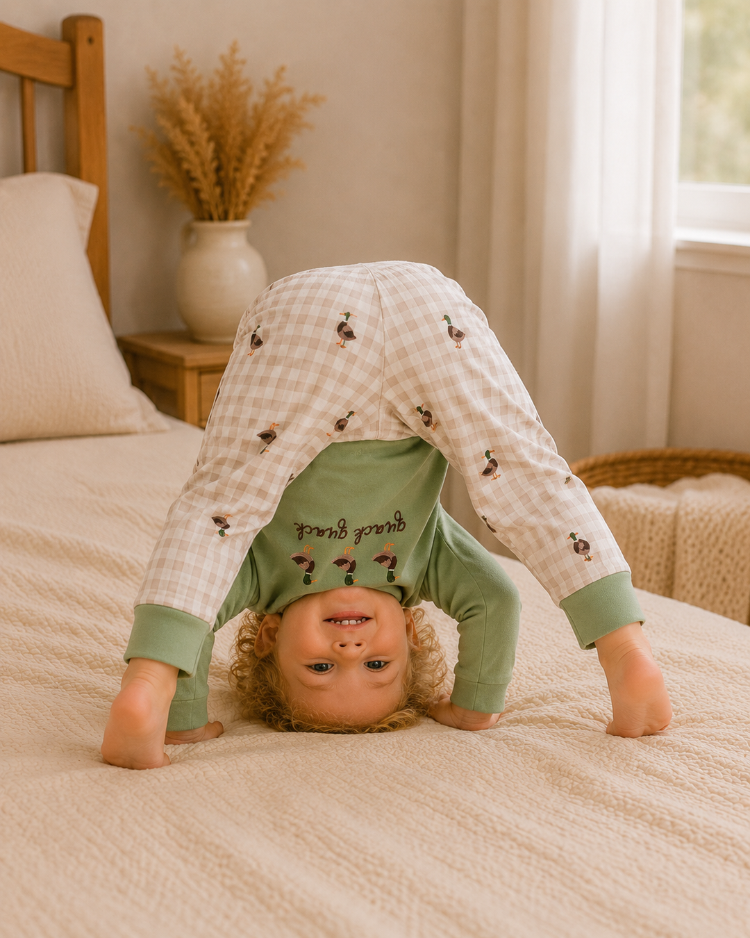 A toddler wearing a green long-sleeve shirt and beige-and-white gingham pants with small printed motifs, performing a handstand on a cream-colored bed in a softly lit bedroom.