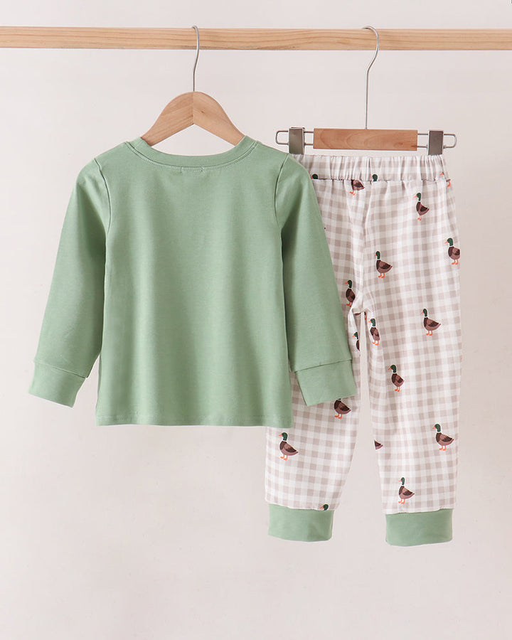 A two-piece pant set hanging on wooden hangers against a plain light beige wall. The set includes a sage green long-sleeve top and white gingham pants with a small duck print and sage green cuffs.