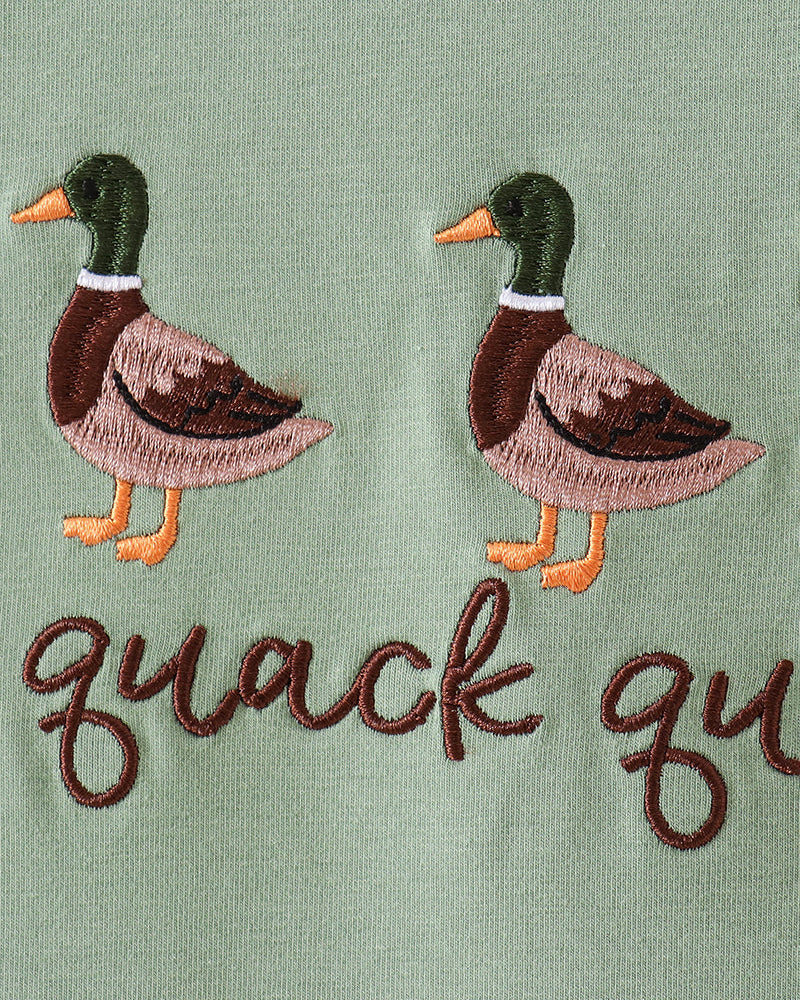 Close-up of embroidered ducks and the words "quack qu" on a sage green fabric surface.