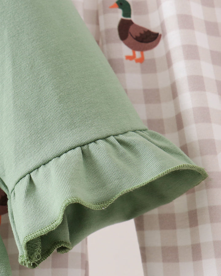 Close-up of a sage green fabric product with a ruffled hem, laid over a beige and white gingham patterned fabric featuring duck illustrations.