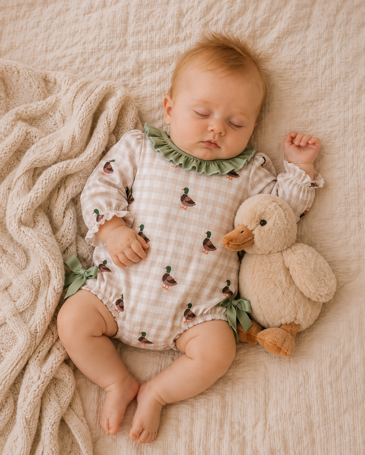 A sleeping baby wearing a beige and white gingham romper with green ruffle trim and duck prints, lying on a textured cream blanket next to a plush duck toy.