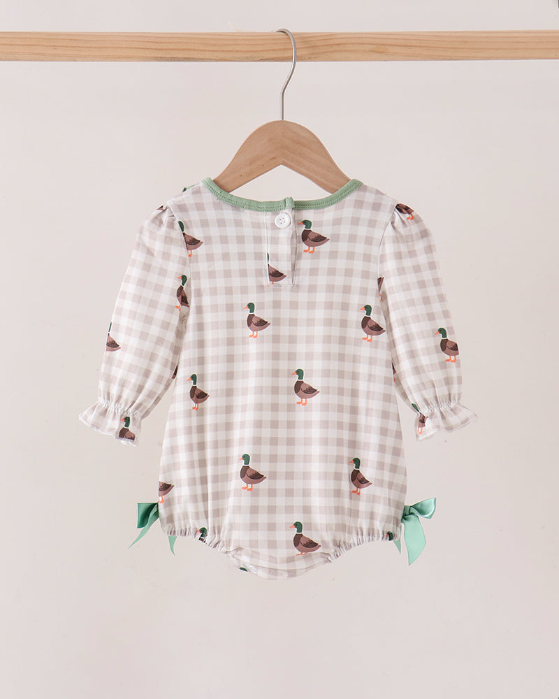 A baby romper with a beige and white gingham pattern, decorated with small green and brown duck prints, hanging on a wooden hanger against a light beige wall.