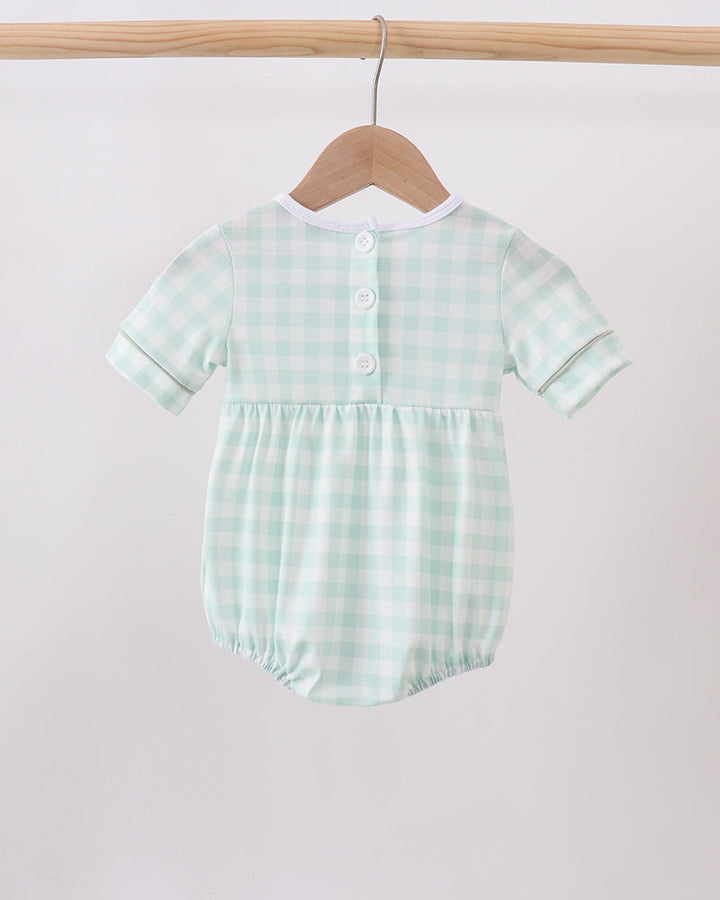 A baby romper in a light mint green and white gingham pattern, hanging on a wooden hanger from a wooden clothes rack against a plain light background.