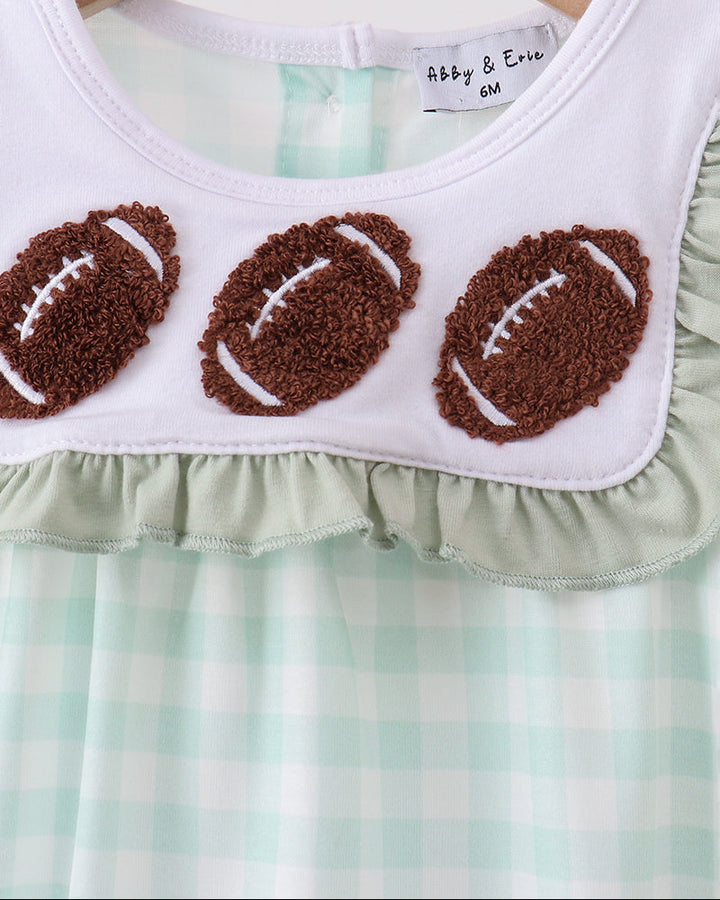 A close-up of a baby or toddler dress hanging on a wooden hanger. The top portion is white with three brown textured football appliqués outlined in white, and the lower portion is mint green and white gingham.