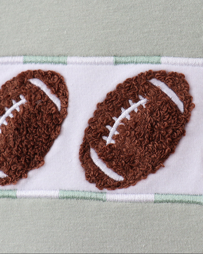 Close-up of a white fabric band with two brown football appliqués and thin mint green stripes, set against a pale sage green fabric background.