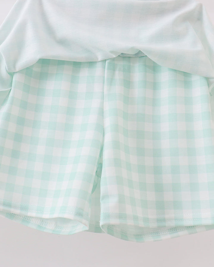 A close-up of a mint green and white gingham check patterned garment, likely shorts or a skirt, with a solid mint green top draped over the waist, set against a plain light background.
