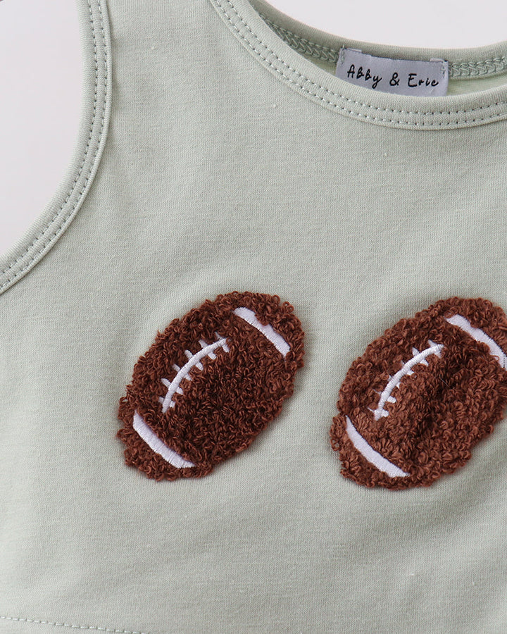 Close-up of a sleeveless baby top in pale sage green, featuring two brown textured football appliqués with white laces on the chest. The inner neck label reads Abby &amp; Eve.