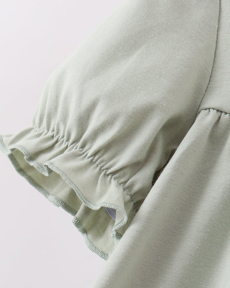 Close-up of a light sage green garment with a gathered ruffled sleeve detail, showing the soft fabric texture against a plain light background.