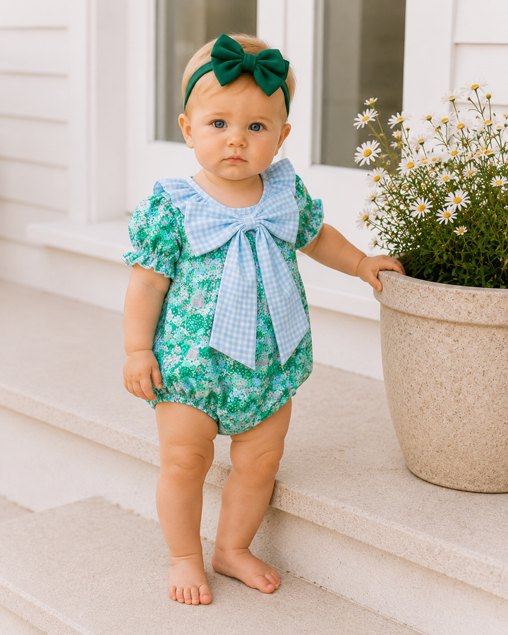 Baby in a green floral outfit with a blue bow standing on steps next to a potted plant.
