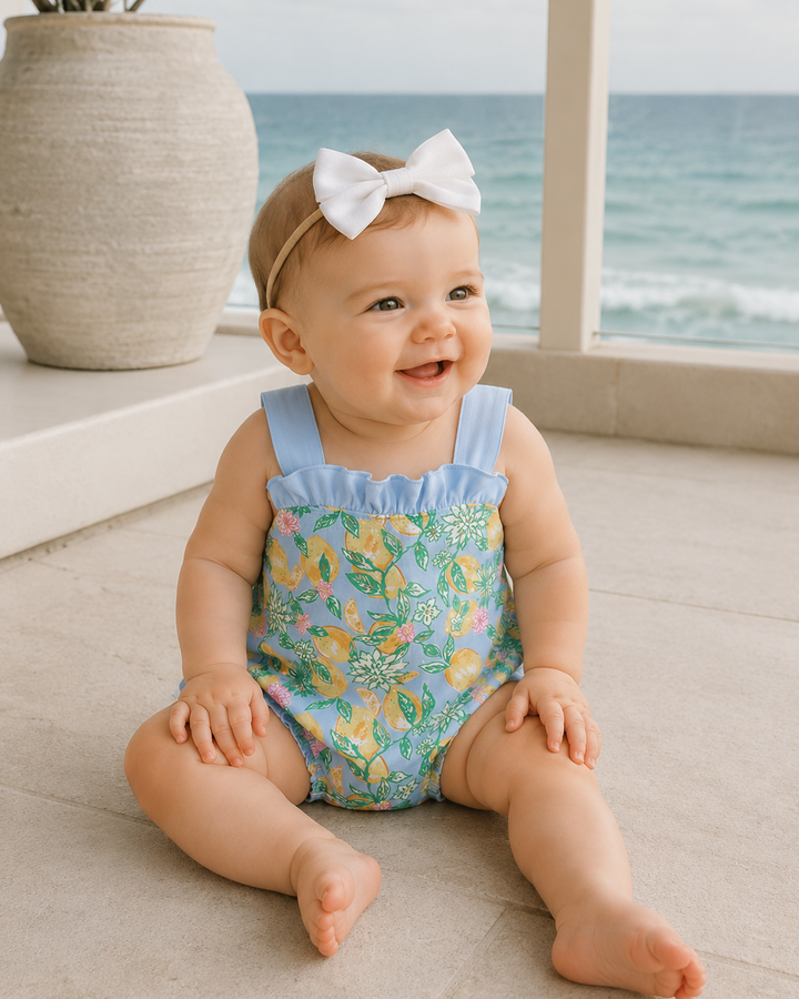 Baby sitting on patio with a floral romper and white bow, ocean view in the background