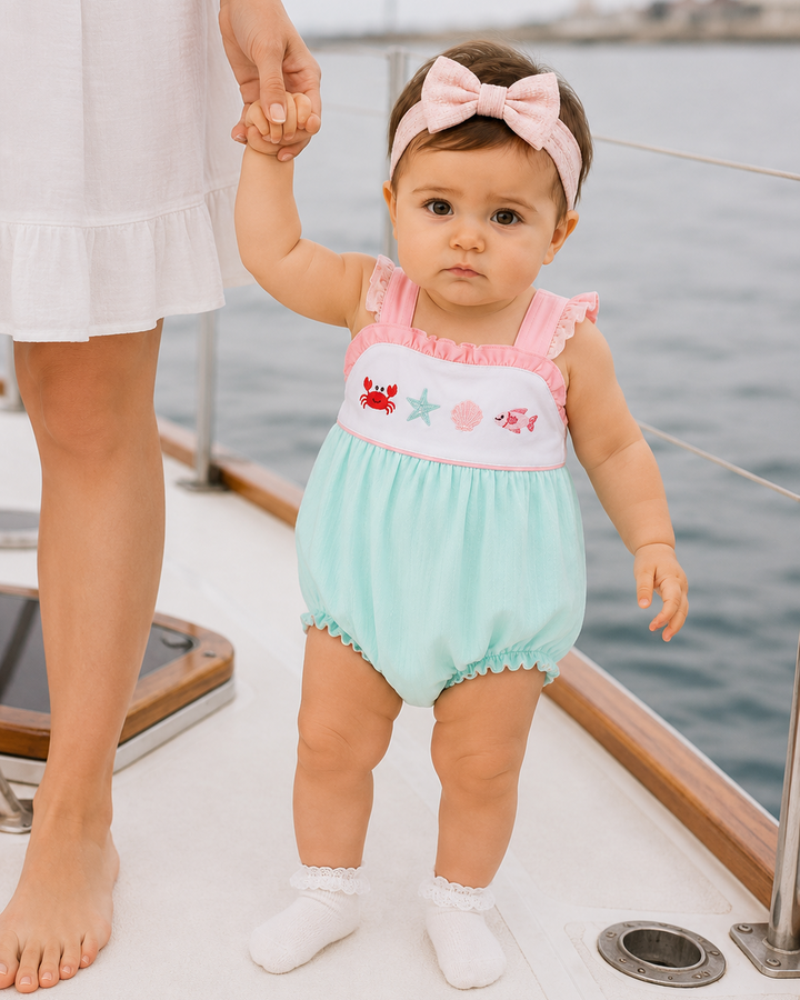 Baby in a light blue outfit with pink accents standing on a boat holding hands with an adult.