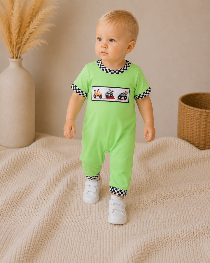 A baby standing on a beige knit blanket wearing a bright lime green footless one-piece jumpsuit with black and white checkered trim on the collar, sleeve cuffs, and ankle cuffs. The chest features a rectangular graphic with small red, white, and black tractors.