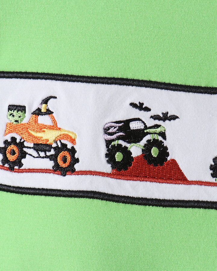 Close-up of a bright green garment featuring a horizontal embroidered band with Halloween vehicles, including an orange ATV with a green skull driver and a black monster truck on a red ramp.