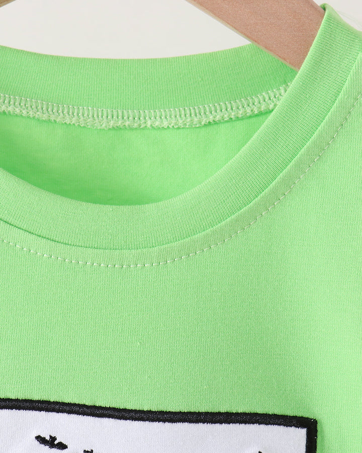 Close-up of a bright lime green crewneck t-shirt hanging on a wooden hanger, showing the ribbed collar and a partial white graphic patch with black trim at the bottom.