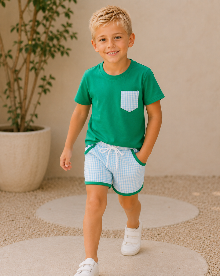 Child wearing a green shirt and checkered shorts standing in front of a plant.