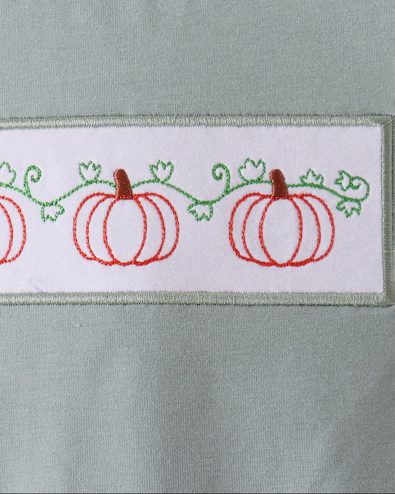 Close-up of a sage green crewneck shirt featuring a white rectangular chest patch with embroidered red pumpkins and green vines.