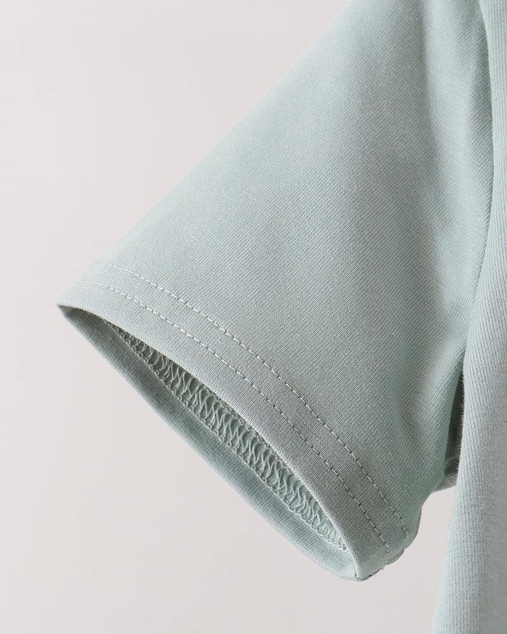 Close-up of a light sage green short-sleeve t-shirt showing the sleeve cuff with neat double stitching, set against a plain off-white background.