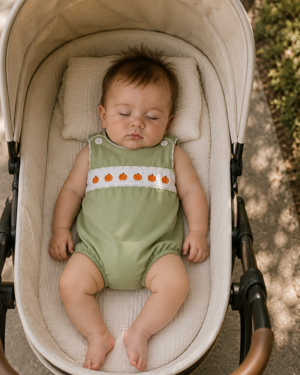 A baby wearing a sage green sleeveless romper with a white chest band and orange pumpkin embroidery, sleeping inside a cream-colored stroller outdoors.