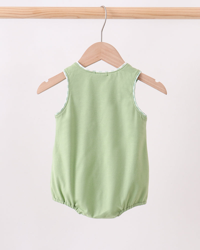 A light sage green sleeveless baby romper with elasticated leg openings, hanging on a wooden hanger from a wooden clothes rack against a plain off-white background.