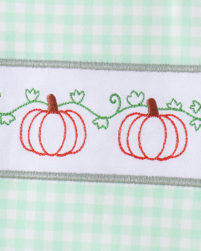 A close-up of a white embroidered band featuring two orange pumpkins with green vines and leaves, bordered by a gray stitched edge, set against a mint green and white gingham background.