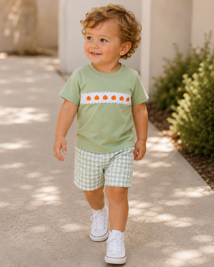 A toddler walking outdoors on a sunlit concrete path, wearing a light sage green short-sleeve t-shirt with a horizontal white stripe featuring orange pumpkin motifs, paired with green-and-white gingham shorts and white sneakers.