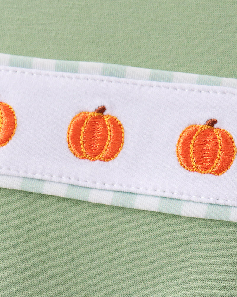 A close-up of a white fabric trim featuring embroidered orange pumpkins with brown stems, bordered by a light green and white gingham pattern, set against a light sage green background.