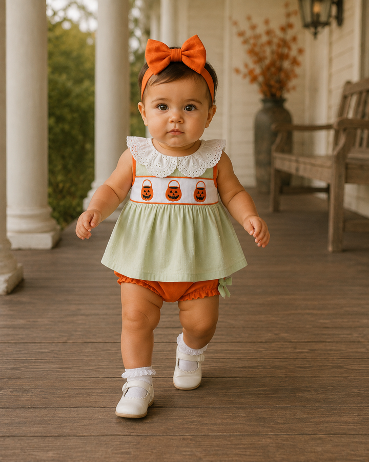 A toddler girl walking on a wooden porch wearing a light green sleeveless dress with a white lace collar and three orange pumpkin motifs across the chest, paired with orange bloomers, a matching orange bow headband, white socks, and white shoes.