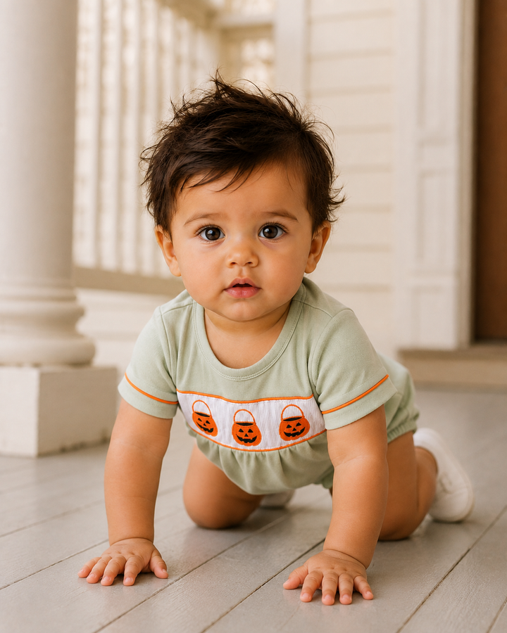 A baby crawling on a light-colored wooden porch wearing a sage green short-sleeve romper with a white chest panel featuring three embroidered orange jack-o&