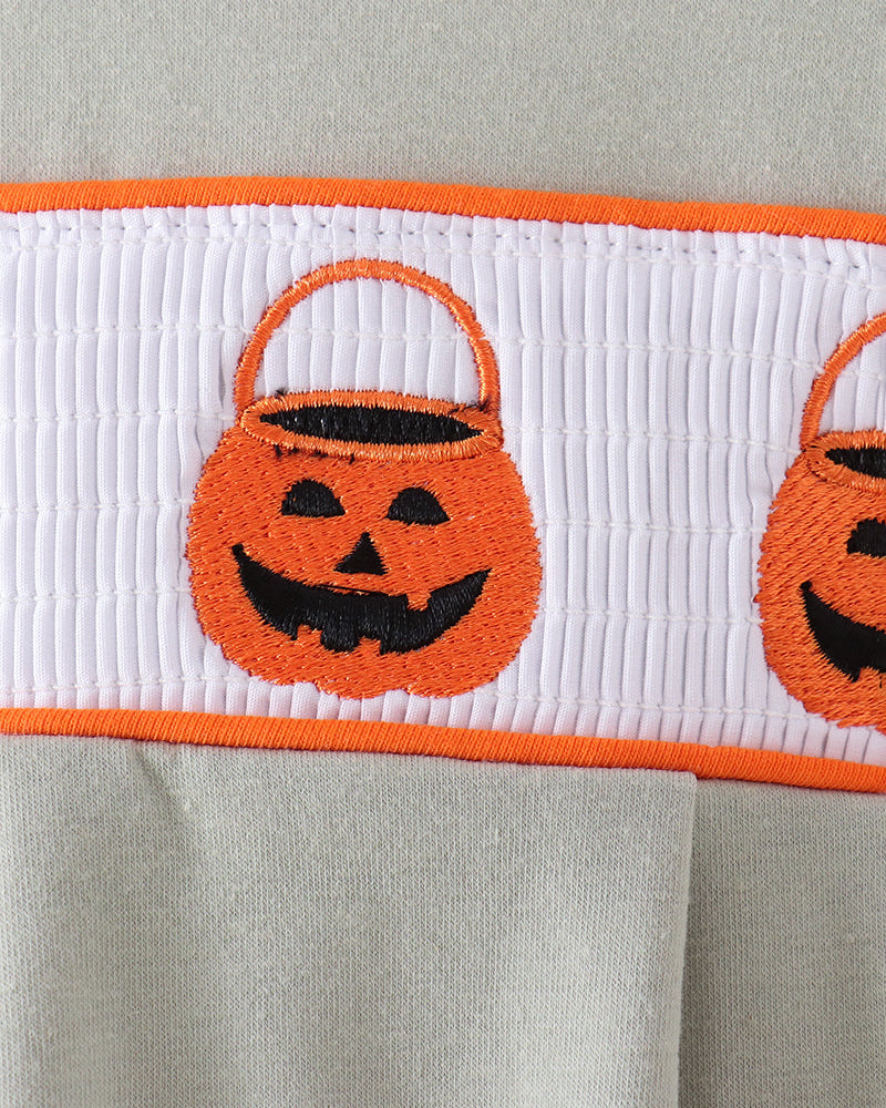 Close-up of a light gray garment featuring a white horizontal band with orange trim and embroidered orange pumpkin bucket motifs with black facial details.