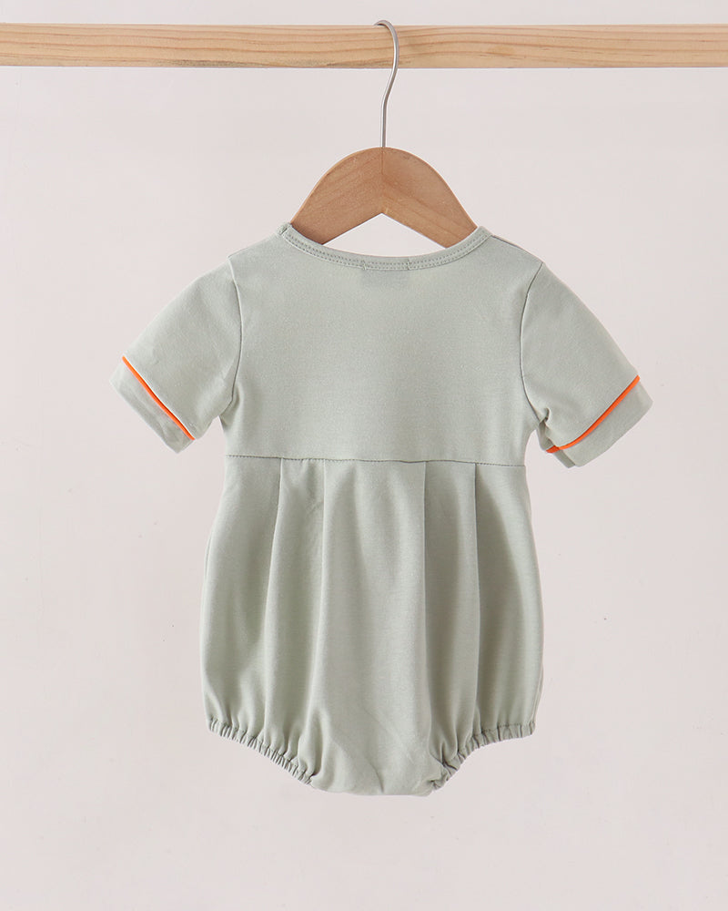 A baby romper in sage green with orange piping on the sleeve cuffs, hanging on a wooden hanger from a wooden clothes rack against a light beige background.