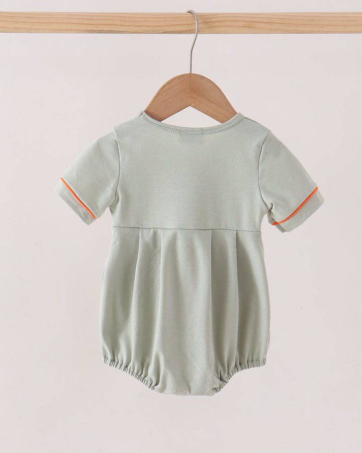 A baby romper in sage green with orange piping on the sleeve cuffs, hanging on a wooden hanger from a wooden clothes rack against a light beige background.