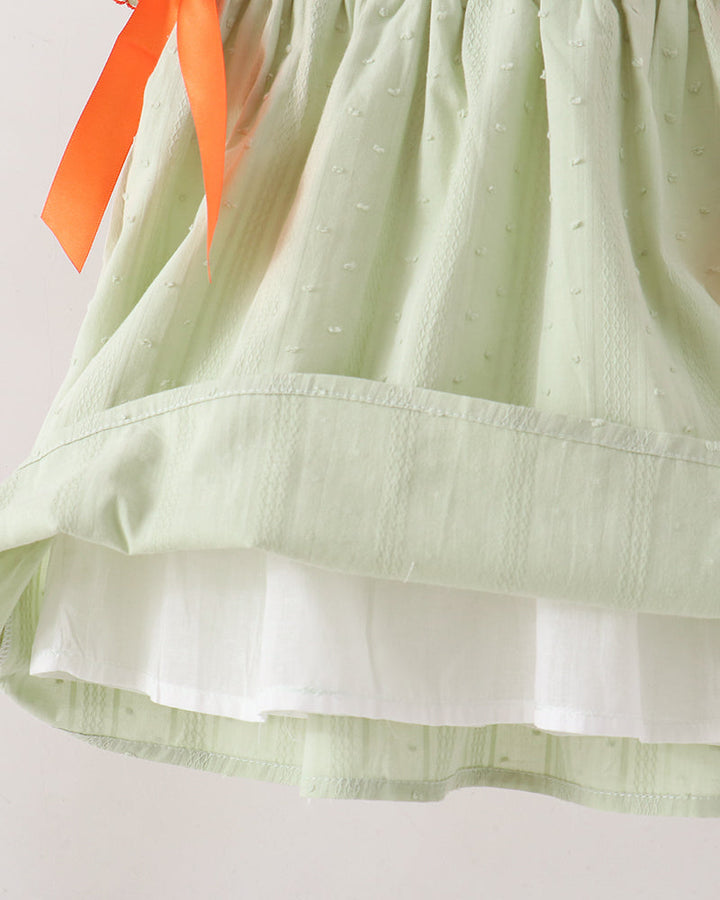 Close-up of a light sage green dress with a textured eyelet fabric, a white underlayer visible at the hem, and an orange ribbon tied near the top.