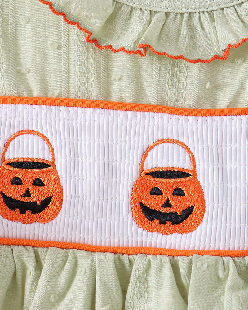 Close-up of a light sage green dress with orange trim, featuring a white smocked band decorated with two embroidered orange pumpkin bucket motifs.