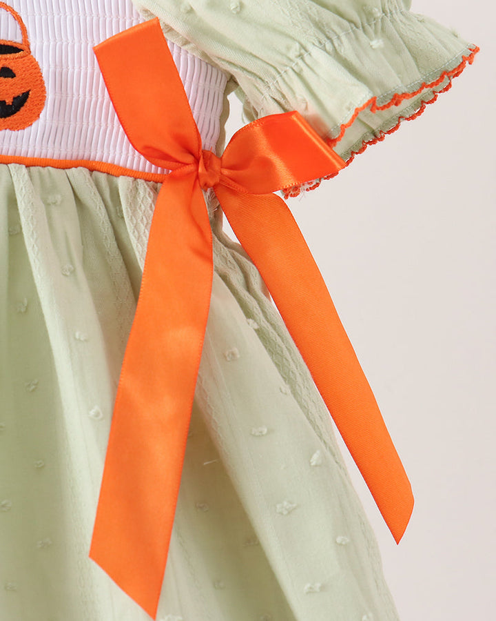Close-up of a light sage green dress with a white and orange striped waistband featuring an embroidered orange pumpkin, tied with a large orange satin ribbon bow.
