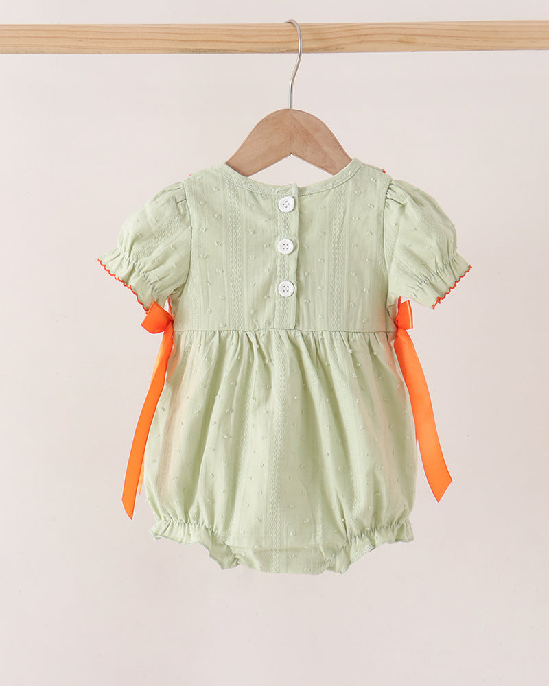 A light sage green baby romper with short puff sleeves, three white buttons on the back, and bright orange ribbon bows on the sides, hanging on a wooden hanger against a plain beige wall.