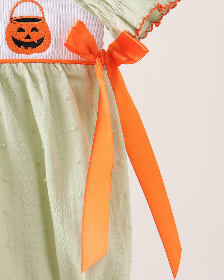 A close-up of a light sage green baby dress with a white and orange embroidered pumpkin bucket band, featuring a long orange ribbon tied in a bow on the hip.