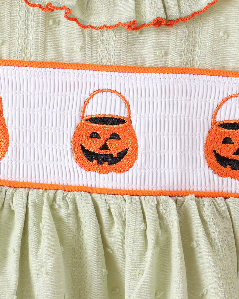 Close-up of a baby dress with a pale sage green textured fabric, featuring an orange and white smocked band with embroidered jack-o&