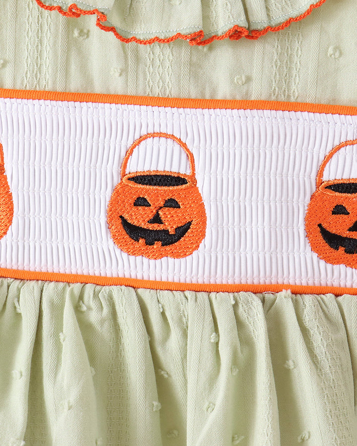 Close-up of a baby dress with a pale sage green textured fabric, featuring an orange and white smocked band with embroidered jack-o&