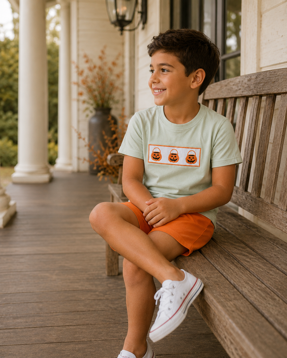 A young boy sits on a wooden porch bench wearing a light sage green t-shirt with a graphic of three orange jack-o'-lantern buckets, orange shorts, and white sneakers, looking to the side with a smile.