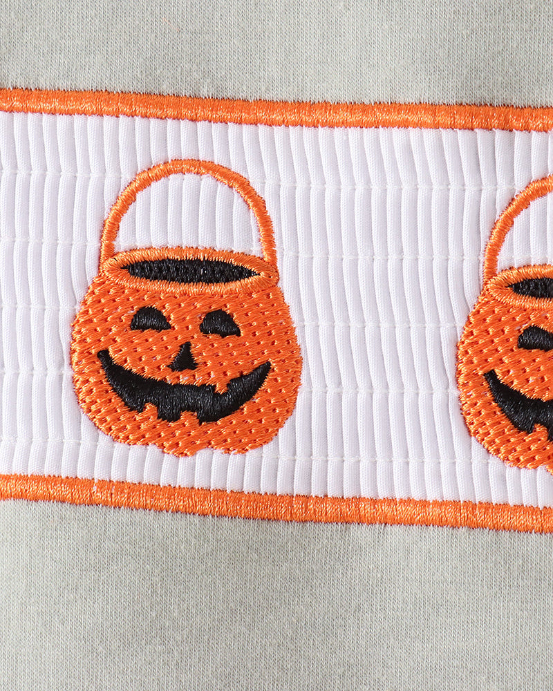 Close-up of a white ribbed fabric trim featuring orange embroidered borders and two orange pumpkin bucket motifs with black stitched faces.