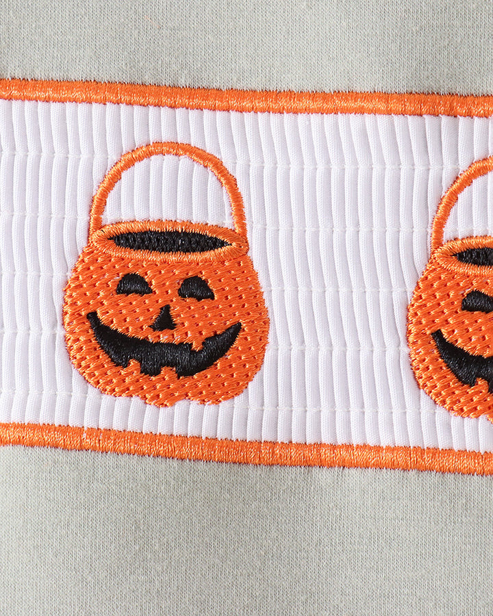 Close-up of a white ribbed fabric trim featuring orange embroidered borders and two orange pumpkin bucket motifs with black stitched faces.