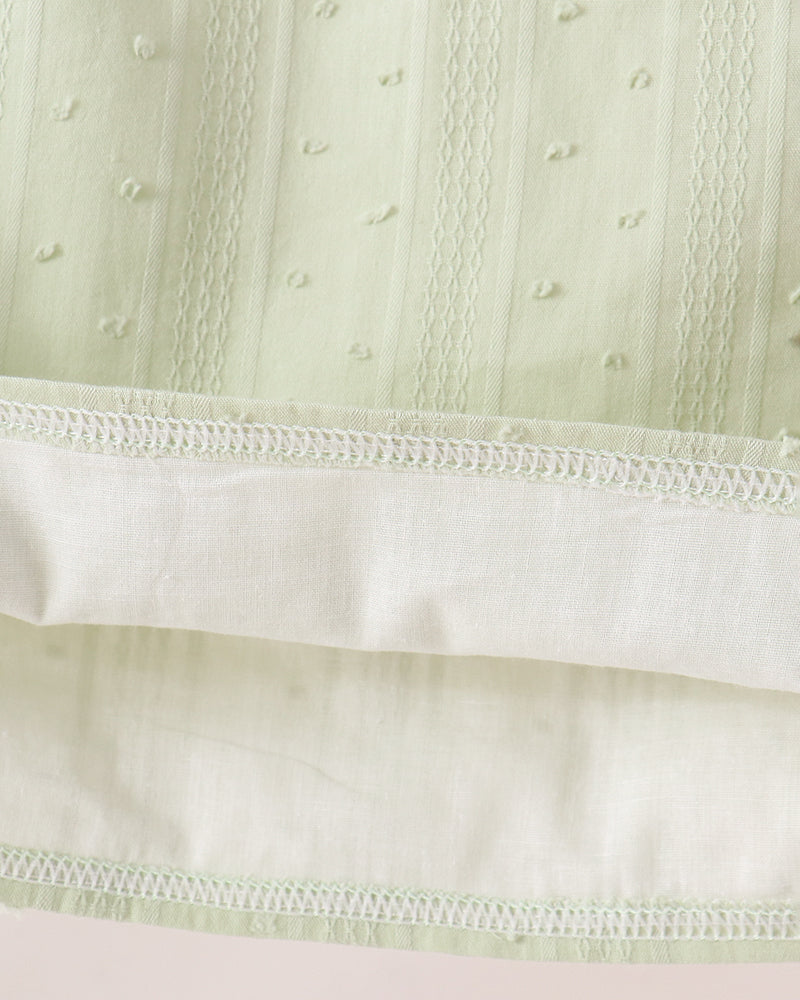 Close-up of a pale sage green embroidered fabric with vertical stitched details and a contrasting off-white hemmed edge.