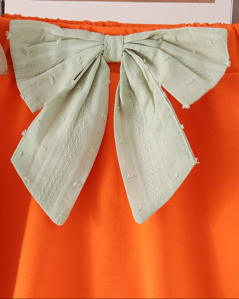 A close-up of a bright orange garment featuring a large pale mint green bow with textured fabric and small pom-pom details, hanging on a wooden hanger.