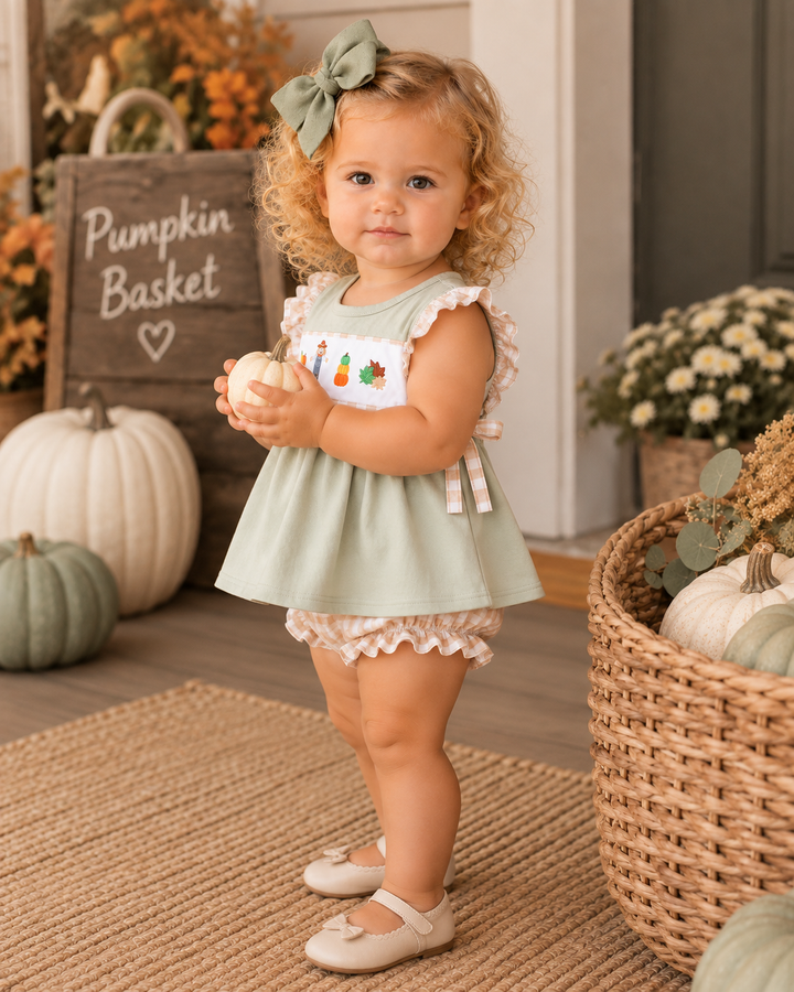 A toddler girl stands in a fall-themed nursery or porch setting wearing a sage green sleeveless dress with ruffled trim and a small embroidered patch, paired with matching ruffled shorts and beige shoes. Pumpkins, a basket, flowers, and a sign reading &