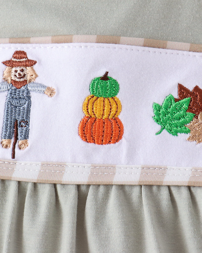 Close-up of a baby dress featuring a white horizontal band with embroidered autumn motifs, including a scarecrow, stacked pumpkins, and fall leaves, in sage green and beige.