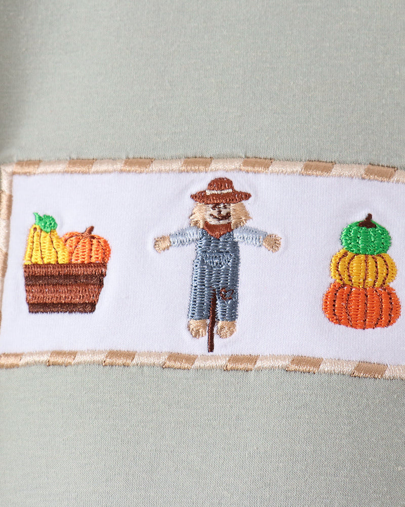 Close-up of a sage green garment featuring a white rectangular embroidered patch with a scarecrow in the center and two stacked pumpkins on either side.